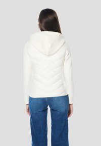 Fehér steppelt dzseki kapucnival, testhezálló fazonban és sima ujjakkal. Hátulról, szürke háttér előtt látható.