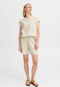 Top beige chiaro a maniche corte con scollatura a V e vita elastica, abbinato a pantaloni corti coordinati. Tessuto morbido e testurizzato. Sandali marroni.