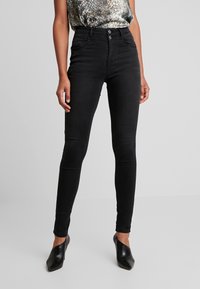 Jeans ajustados de talle alto negros con una textura suave, cierre de dos botones y cuatro bolsillos, combinados con una blusa estampada y tacones negros.