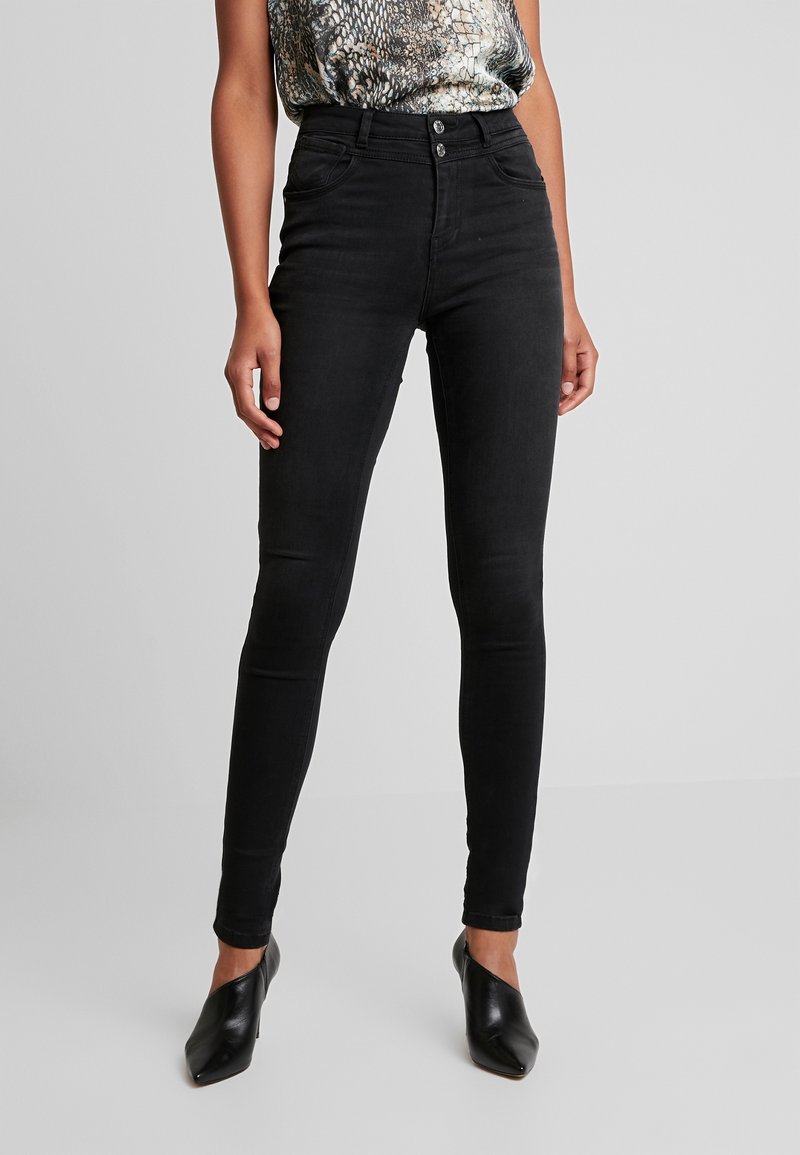 Jeans ajustados de talle alto negros con una textura suave, cierre de dos botones y cuatro bolsillos, combinados con una blusa estampada y tacones negros.