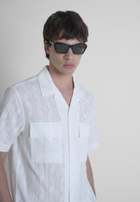 Camicia bianca a maniche corte con un motivo geometrico testurizzato e due tasche sul petto. Indossata con occhiali da sole neri angolari.