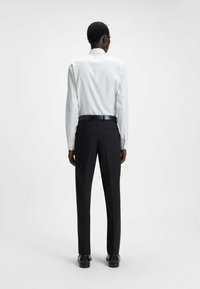 Camicia bianca a maniche lunghe con un piccolo motivo ripetuto, abbinata a pantaloni neri su misura e scarpe nere, vista da dietro.