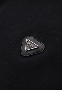 Černá tkanina s hladkým povrchem, na níž se nachází trojúhelníková kožená nášivka v černé barvě s nápisem "OMBRE" vyraženým ve stříbrném písmu.