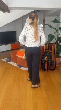 Femme aux longs cheveux attachés en queue de cheval se tient dans un salon avec un sol en bois, un canapé orange, une chaise pliante en bois et une grande plante en pot.