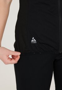 Svart atletisk ytterlager med en slät textur, zip-stängning och en "WX-TECH"-logga vid fållen, med en närbild av en hand som greppar tyget.