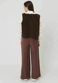 Gilet en polaire marron à col haut, porté sur une blouse blanche à fleurs. Associé à un pantalon ample marron foncé et des chaussures blanches.