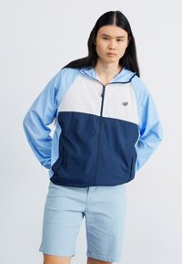Veste zipée légère en bleu et marine, présentant un design coloré avec une capuche et des poches avant. Texture lisse, détails minimaux.