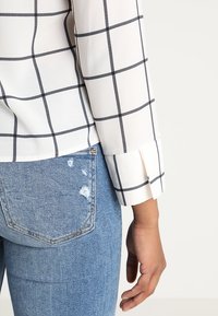 Camicia bianca con motivo a griglia nera, dotata di maniche con polsini, indossata sopra jeans blu strappati con dettagli sulle tasche.