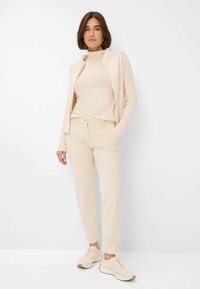 Beige stretch top met lange mouwen, mouwloze lichte jas en taps toelopende lichtbeige broeken. Casual sneakers maken de outfit compleet. Effen achtergrond.