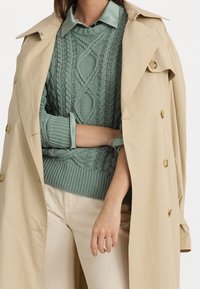 Beige trenchcoat med guldfärgade knappar över en mintgrön kabelstickad tröja. Ljusblå krage synlig. Krämfärgade byxor under.