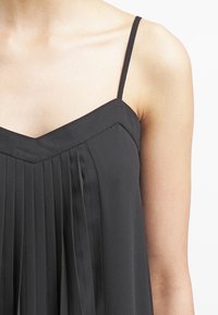 Svart ärmlös topp med v-ringning, med vertikala veck och tunna justerbara axelband gjorda av ett mjukt och lätt tyg.