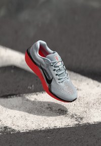 Baskets de sport gris clair avec des accents noirs, semelle rouge, dessus en maille texturée et design à lacets, montrées en plein air sur un fond sombre.