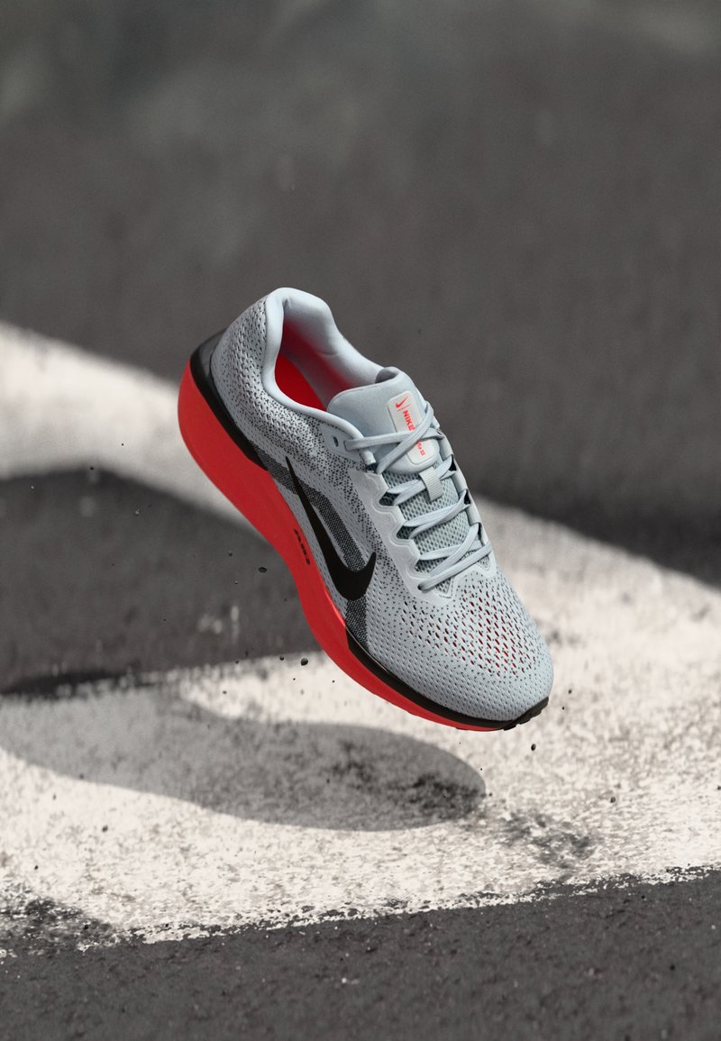 Baskets de sport gris clair avec des accents noirs, semelle rouge, dessus en maille texturée et design à lacets, montrées en plein air sur un fond sombre.