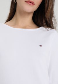 Witte shirt met lange mouwen, een brede halslijn en een geribbelde structuur, met een klein rood, wit en blauw geborduurd logo op de borst.