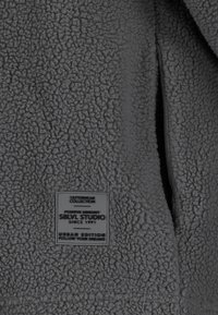 Grau gefütterte Jacke mit einer strukturierten Oberfläche, seitlicher Reißverschlusstasche und einem Markenetikett mit Details zur Kollektion und Auflage.