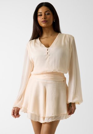 Femme portant un chemisier à manches longues crème clair avec trois boutons et une mini-jupe assortie à couches, sur un fond blanc uni.