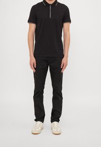 Polo noir avec des finitions blanches, présentant un logo vertical. Associé à un pantalon noir et des baskets claires avec un design texturé.