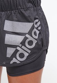 Pantalones cortos deportivos grises con textura de malla, con un destacado logotipo blanco de Adidas en el costado. El diseño incluye una cinturilla elástica.
