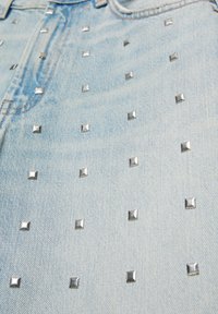 Tissu en denim bleu clair décoré de petits clous carrés argentés disposés en motif quadrillé près d'une couture de poche.