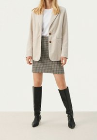 Veste en velours côtelé beige clair à deux boutons, associée à une mini-jupe à carreaux marron et noire, complétée par des bottes noires montant jusqu'aux genoux.