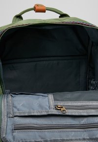 Zelený batoh uvnitř má tmavou podšívku, obsahuje zipovou kapsu a síťovou kapsu. Akcenty zahrnují hnědou koženou klopu a barevný lem.