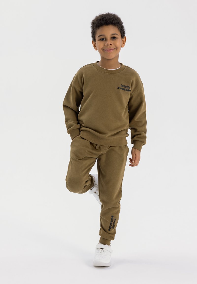 Olijfgroen sweatshirt en broekset van zachte stof; voorzien van de tekst "totally awesome" op de borst en het been; gecombineerd met witte sneakers.