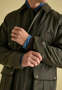 Ciemnozielona ocieplana kurtka z kołnierzem z materiału sztruksowego, zapiętymi kieszeniami i regulowanymi mankietami. Pod nią widoczna jest niebieska koszula dżinsowa.