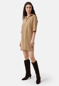 Abito in suede beige con cappuccio, maniche corte e spacchi sui lati, abbinato a stivali neri in suede al ginocchio con punta affusolata.