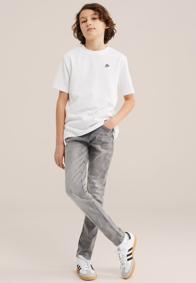 Băiat cu păr creț stă relaxat, purtând un tricou alb, blugi gri și adidași albi cu dungi negre pe un fundal neutru.