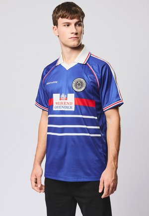 Młody mężczyzna w niebieskiej sportowej koszulce "Weekend Offender" z czerwono-białymi paskami, kołnierzykiem i logo, stojący na tle jednolitego jasnego tła.