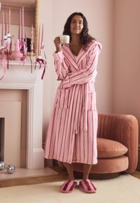 Pink striped robe with a hood and pockets, made of soft fabric; paired with plush pink slippers. Background features a light pink wall and decorative candles.