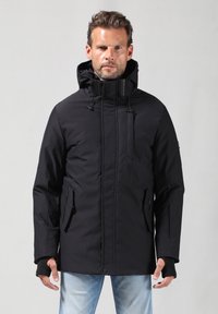 Chaqueta negra aislante con un alto collar, capucha ajustable, bolsillos laterales con cremallera y puños ajustados. Textura suave y material duradero.