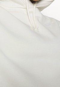 Felpa bianca in morbido tessuto, con cappuccio regolabile e un piccolo logo grigio sul petto. Texture liscia e design minimale.