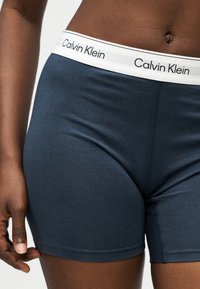 Άτομο με σκουρόχρωμο δέρμα που φοράει μπλε ναυτικό boxer Calvin Klein με λευκή μέση που εμφανίζει το όνομα της μάρκας.