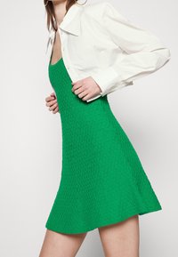 Abito verde con texture e orlo svasato, abbinato a una camicia bianca con colletto, dotata di maniche lunghe e chiusura a bottoni.