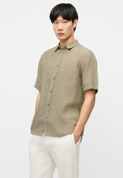 Νέος άνδρας που φοράει μπεζ πουκάμισο με κοντά μανίκια και λευκό παντελόνι, στέκεται με τα χέρια στις τσέπες μπροστά σε μονόχρωμο φόντο.