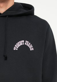 Sort hættetrøje med en afslappet pasform, prydet med et buet "TOMMY JEANS" logo i pink broderi på brystet. Blødt, tekstureret materiale.