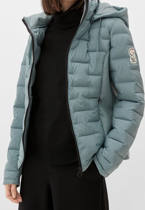Femme portant une veste matelassée à capuche bleu clair sur un pull et un pantalon noirs, avec la veste partiellement dézippée et la main dans la poche.