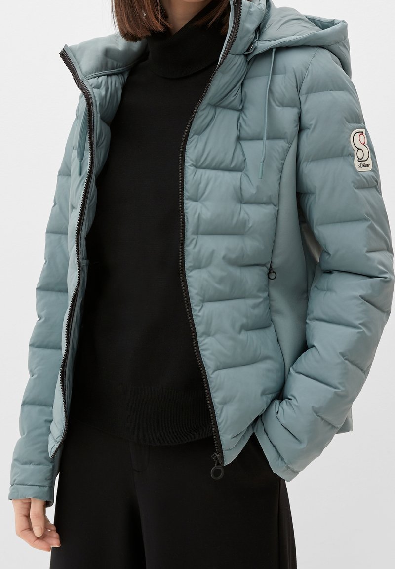Femme portant une veste matelassée à capuche bleu clair sur un pull et un pantalon noirs, avec la veste partiellement dézippée et la main dans la poche.