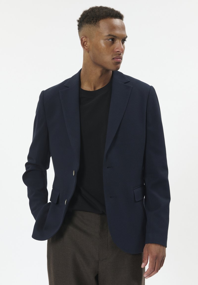 Sötétkék blézer texturált anyagból, egygombos kialakítással, két elülső zsebbel és ezüst gombokkal, fekete környakú pólóval párosítva.