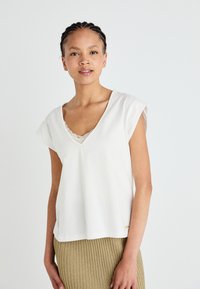 Top bianco a maniche corte con scollatura a V, caratterizzato da un bordo in pizzo sul colletto e una vestibilità comoda, abbinato a una gonna beige a costine.