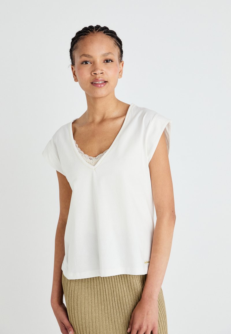 Top bianco a maniche corte con scollatura a V, caratterizzato da un bordo in pizzo sul colletto e una vestibilità comoda, abbinato a una gonna beige a costine.