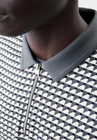 Polo a maniche corte con colletto grigio e mezzo zip, caratterizzato da un motivo triangolare geometrico in bianco e nero su un tessuto testurizzato.