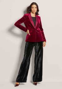 Vrouw in een rood fluwelen blazer, zwarte top, zwarte gestructureerde wijde broek en rode puntige hakken, staand tegen een lichte muur.