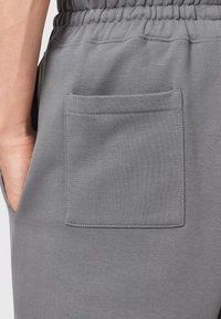 Pantaloni de trening gri, cu o textură netedă, talie elastică și o singură buzunar plat pe partea dreaptă.