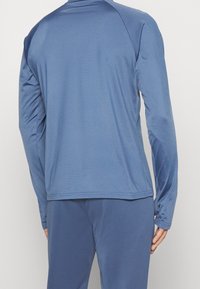 Mies, joka pukeutunut yksiväriseen siniseen pitkähihainen paitaan ja vastaaviin sinisiin housuihin, kuvattu takaapäin yksinkertaisen vaalean taustan edessä.