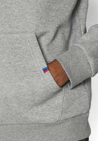 Sudadera gris con textura suave, que cuenta con un bolsillo delantero y una pequeña etiqueta decorativa en rojo y azul en el puño de la manga.