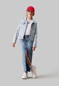 Giacca di jeans, t-shirt bianca con logo, gonna di jeans blu con spacco frontale, cappellino rosso e sneaker bianche. Sfondo neutro.