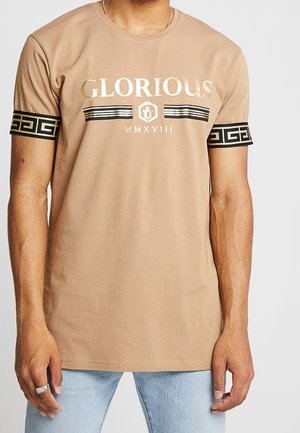Hellbraunes T-Shirt mit dem Schriftzug „GLORIOUS“, schwarze und gold gestreifte Akzente an Ärmeln und Brust, getragen von einer Person mit hellblauen Jeans.