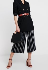 Blazer noir croisé à double boutonnage avec des boutons dorés, ceinture en motif crocodile rouge, culottes rayées, sac à main noir et sandales à talons blocs bordeaux.